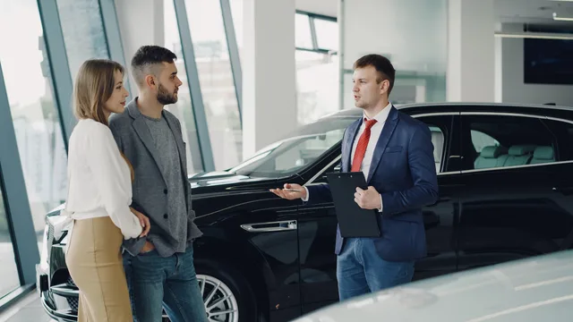 A couple talks with a salesman in a modern car showroom, considering a vehicle purchase. Kundanskaffning som system: mål, kanaler och budget för svenska team